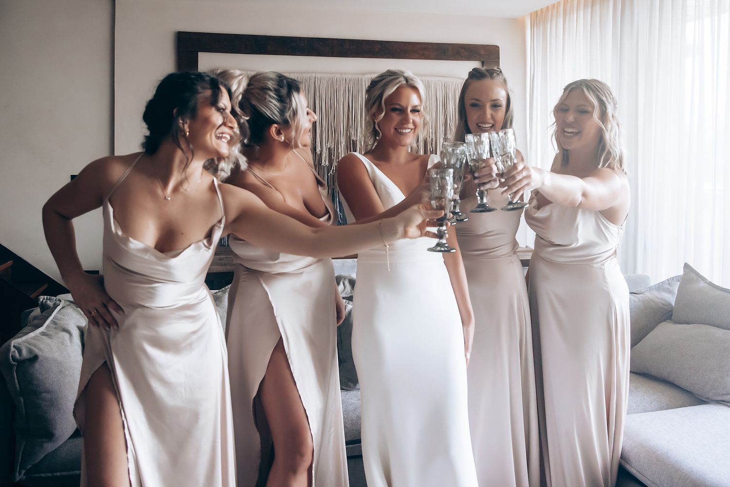 Wedding party in light neutral dresses gathered in sunlit room.