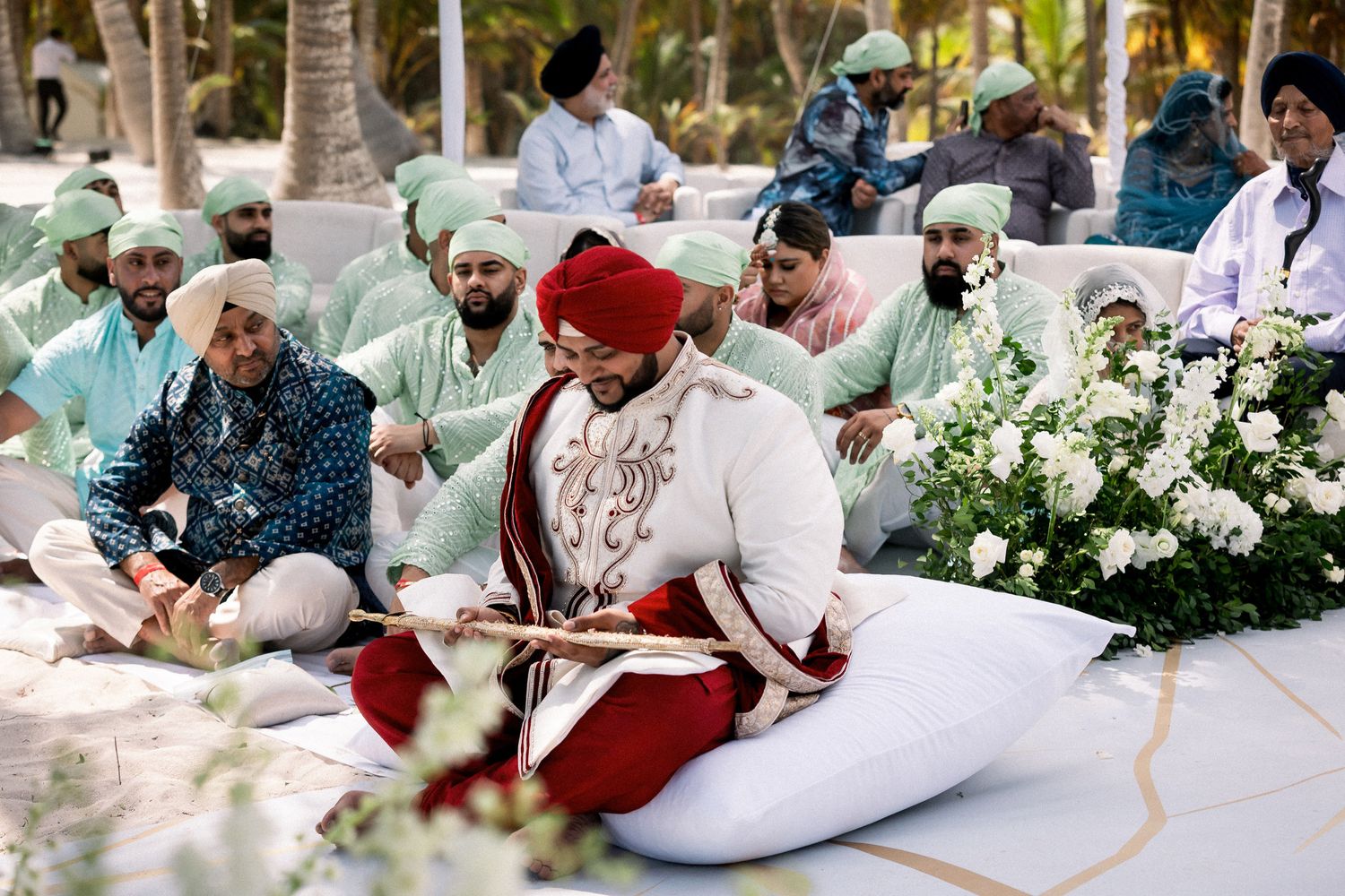 Groom in white sherwani sits during Sikh wedding ceremony with guests in mint turbans.