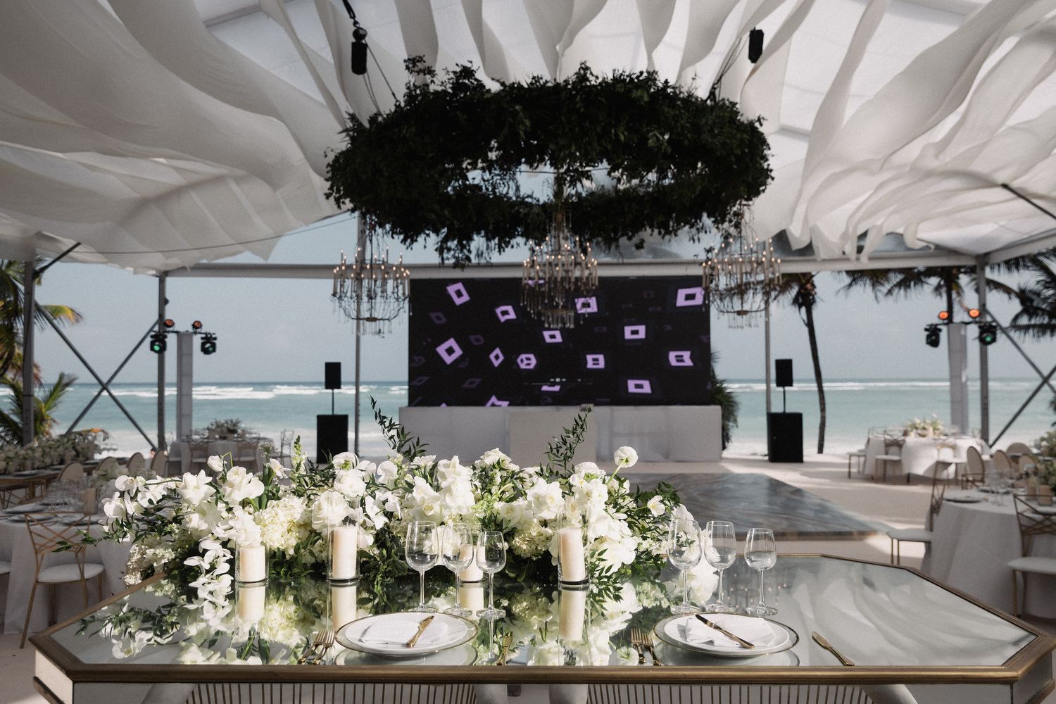 Elegant beachfront wedding reception with white floral centerpieces, candles, and a dark floral chandelier overhead.