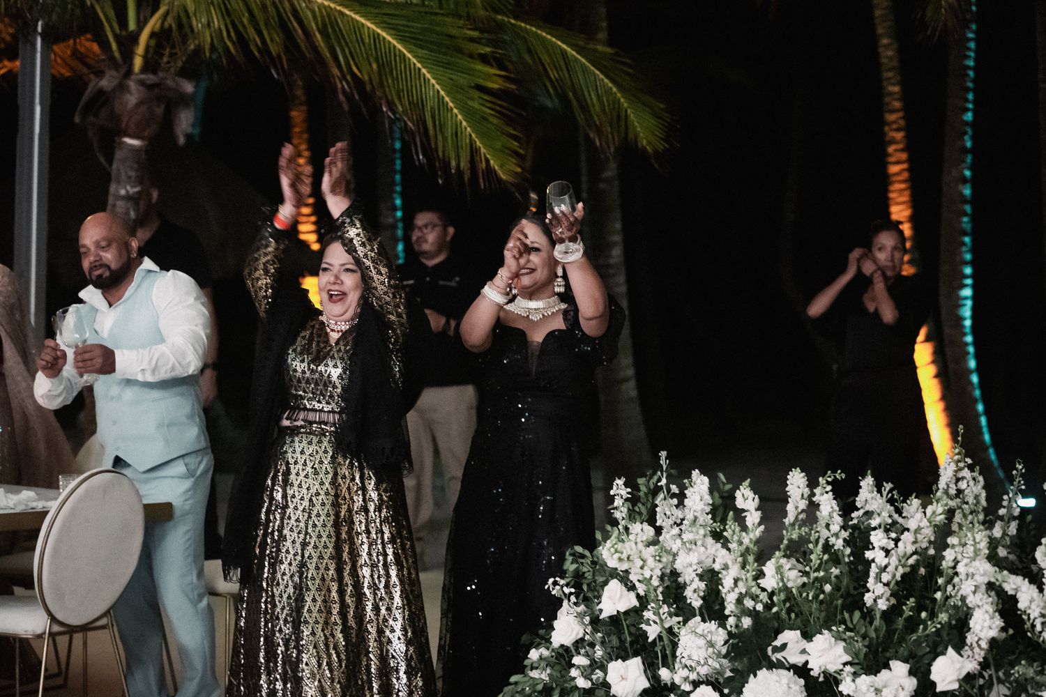 Guests celebrate joyfully at an outdoor wedding reception at night with live drumming and white floral arrangements.