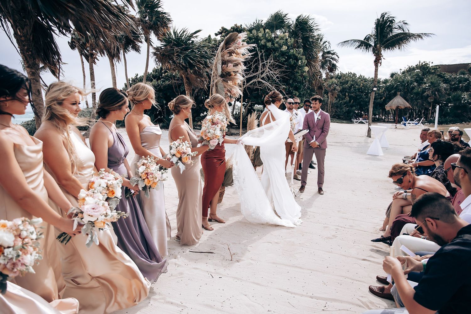 Wedding guests seated in white chairs on beach watching ceremony.