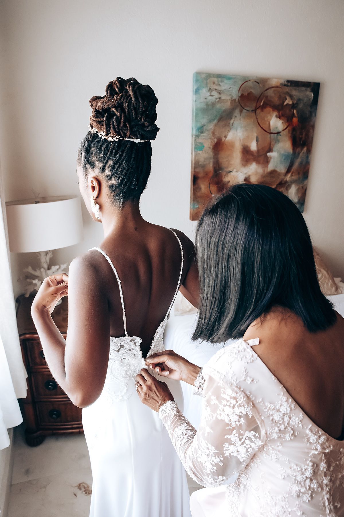 Bridesmaids helping with wedding dress preparations in sunlit room.
