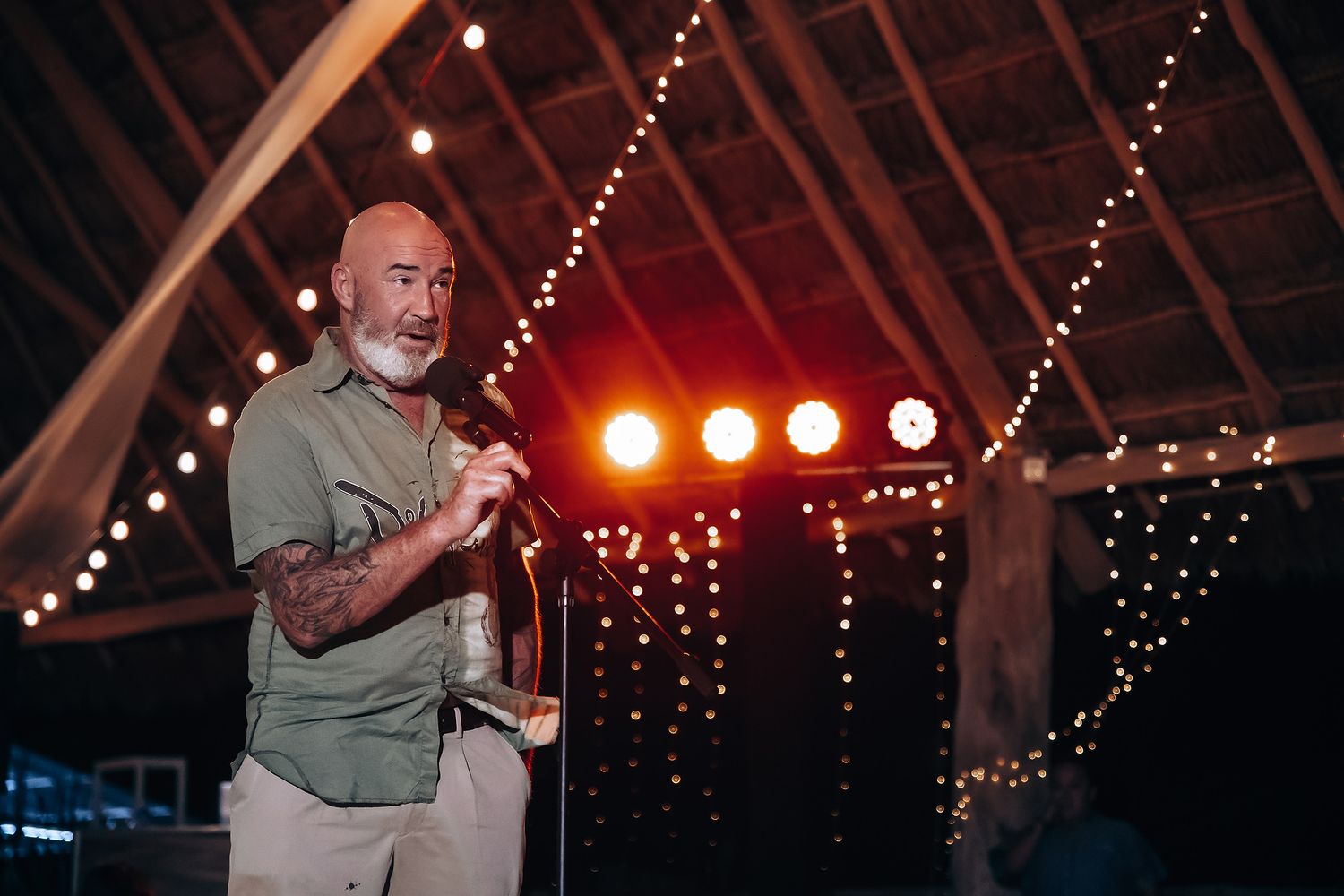 A performer in a green shirt sings on stage under red and white lighting at an indoor venue with string lights.