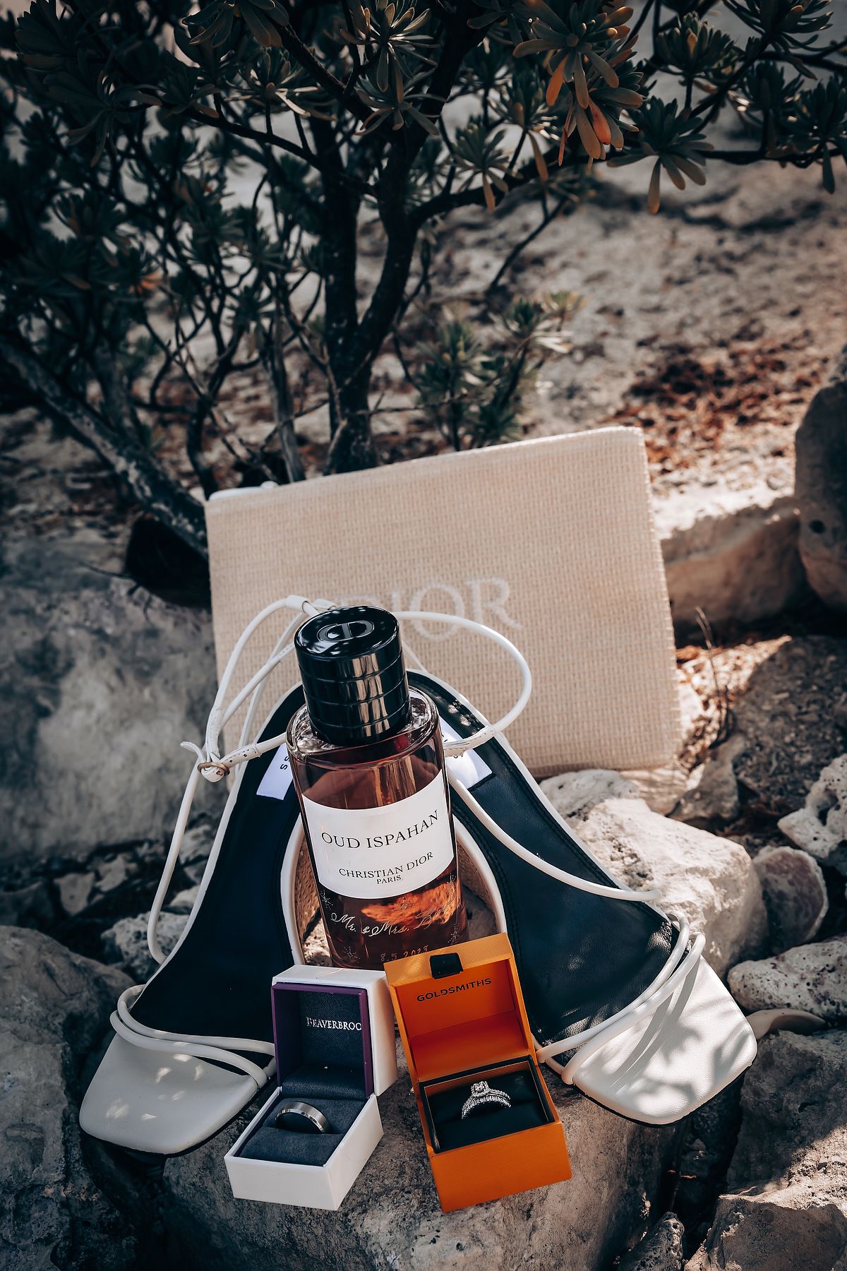 Wedding accessories including perfume bottle, invitation and bridal shoes arranged on textured stone surface in natural light.