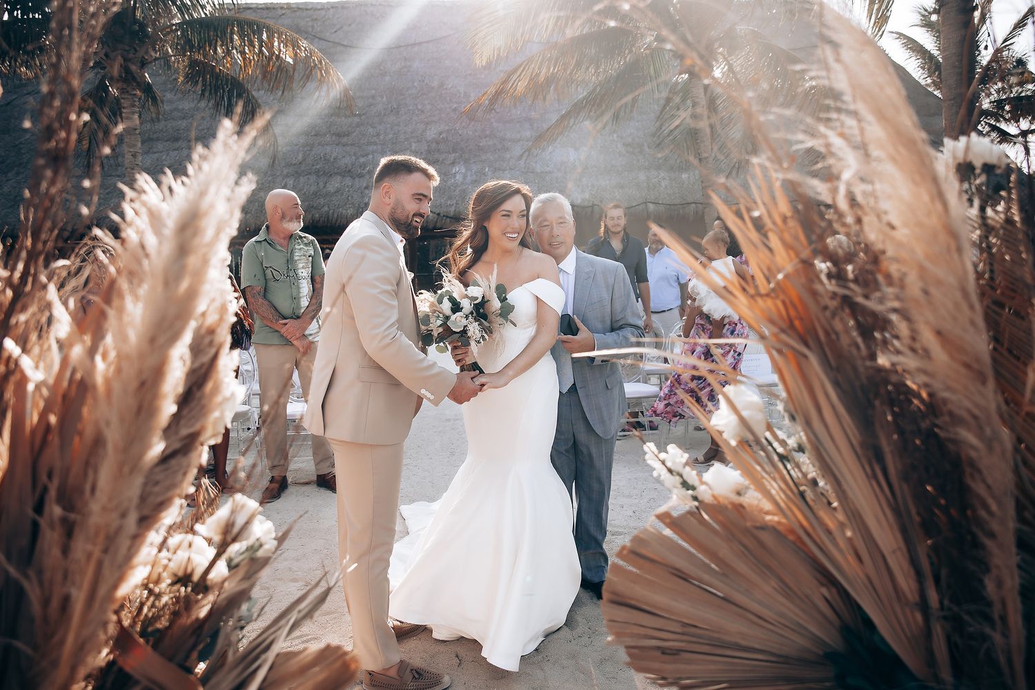 A romantic wedding couple embraces in a sun-drenched garden setting decorated with dried pampas grass arrangements.