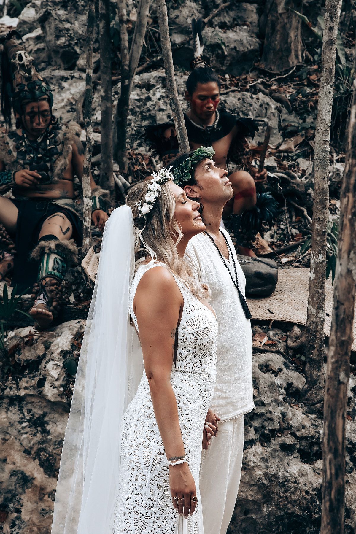 A couple in wedding attire share a candid moment together against a rustic stone wall backdrop in a forest setting.