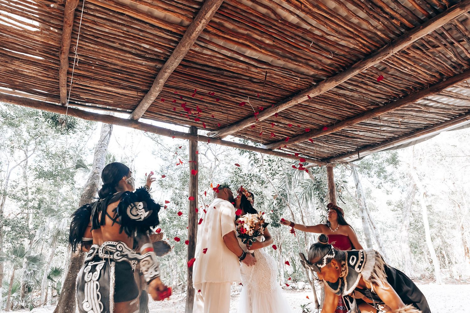 Wedding party dancing beneath a wooden canopy in a natural outdoor setting.