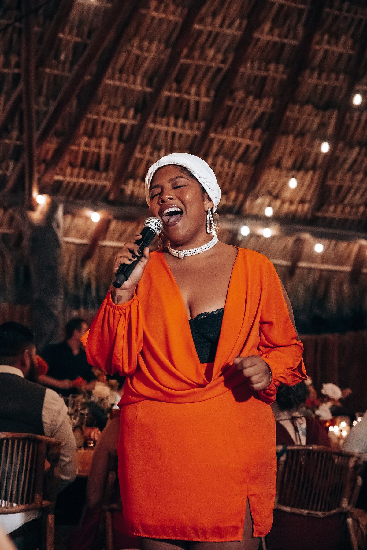 A performer in a bright orange outfit sings passionately on stage under a thatched roof with warm lighting.