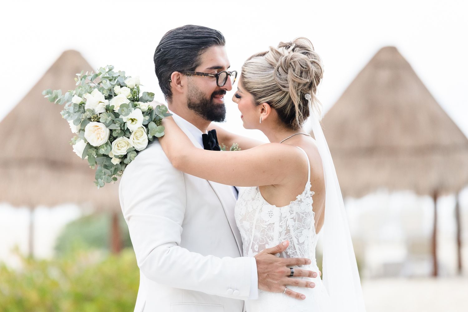 A bride and groom share a romantic moment outdoors at a beach wedding venue, with the bride holding a white bouquet.