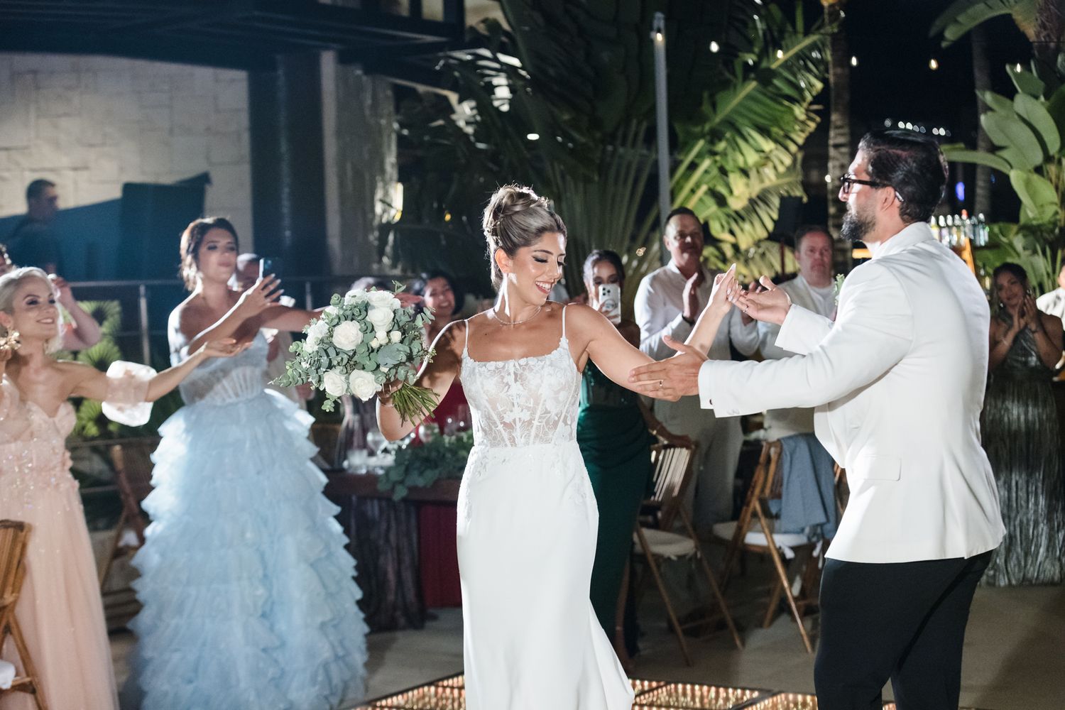 Bride in white gown and groom in white tuxedo celebrate at tropical wedding reception with guests.