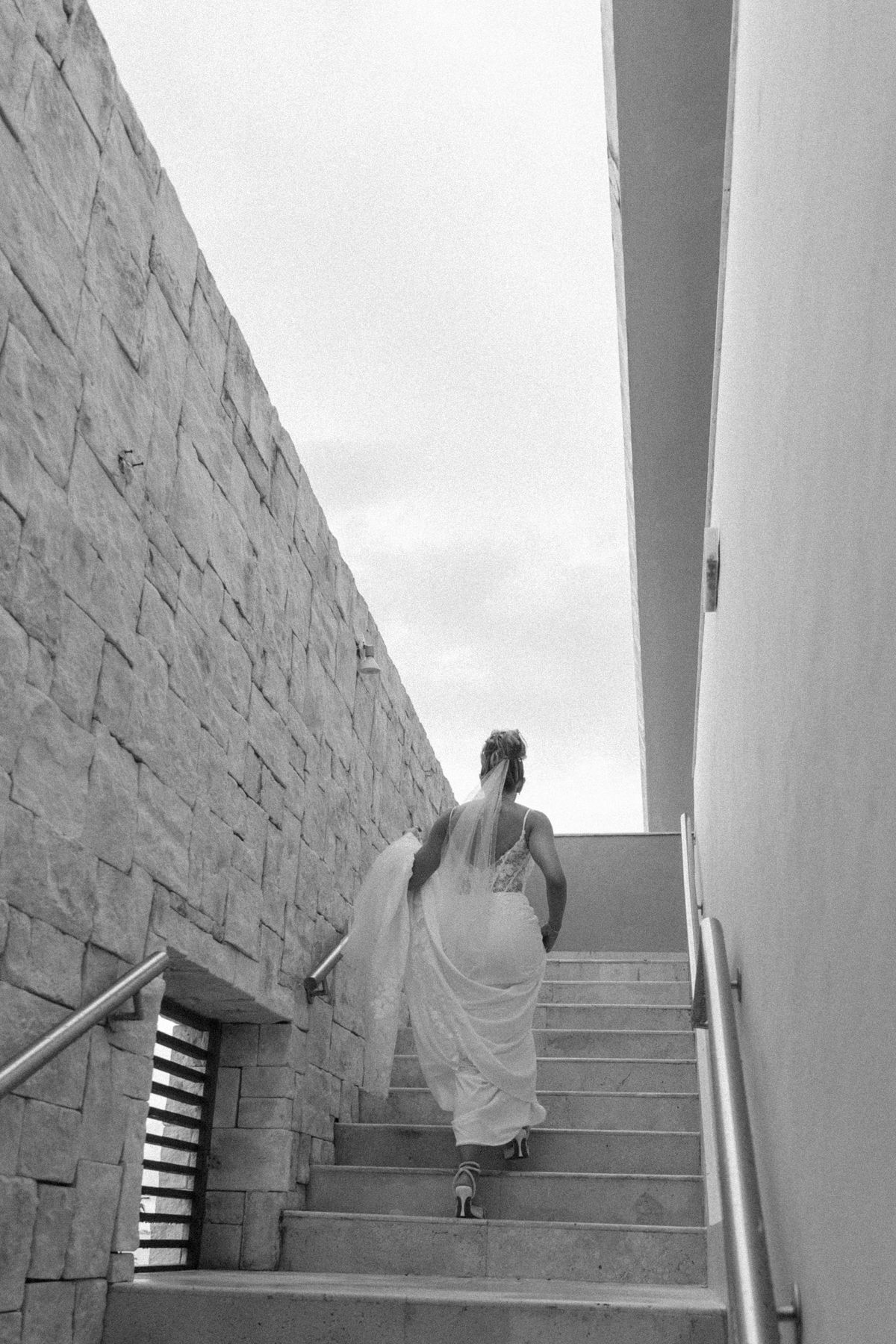 Bride in white wedding gown ascending stone stairs between walls, black and white photo.