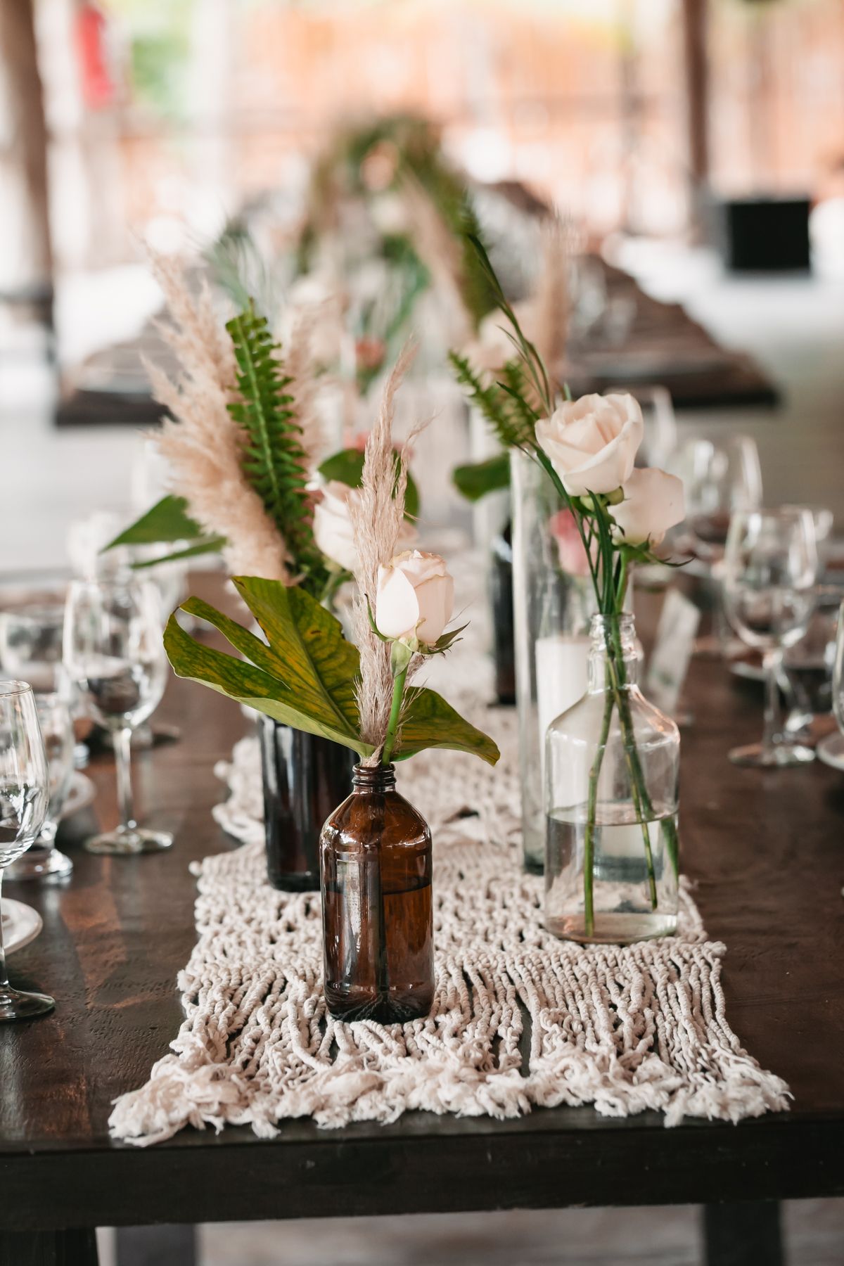 Elegant boho wedding table centerpiece with white roses, tropical leaves, and pampas grass in glass and amber bottles.