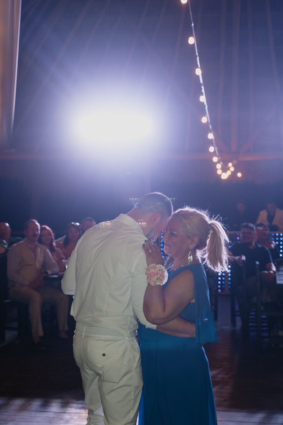 Groom shares a tender mother-son dance under string lights at a tropical wedding reception.