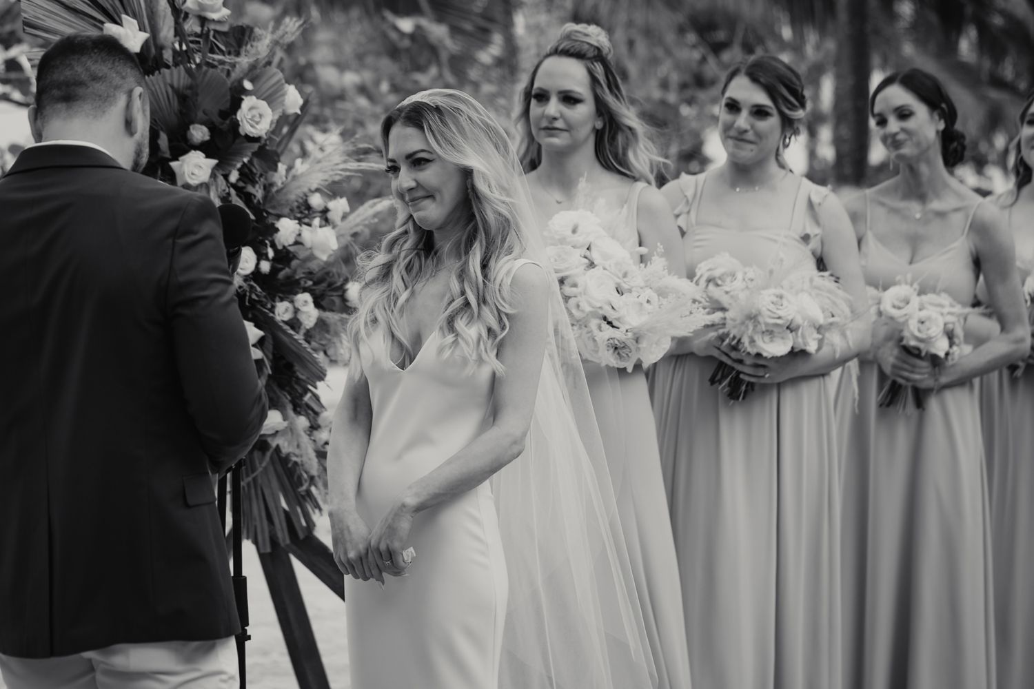Black and white photo of smiling bride at altar with bridesmaids holding white bouquets behind her.