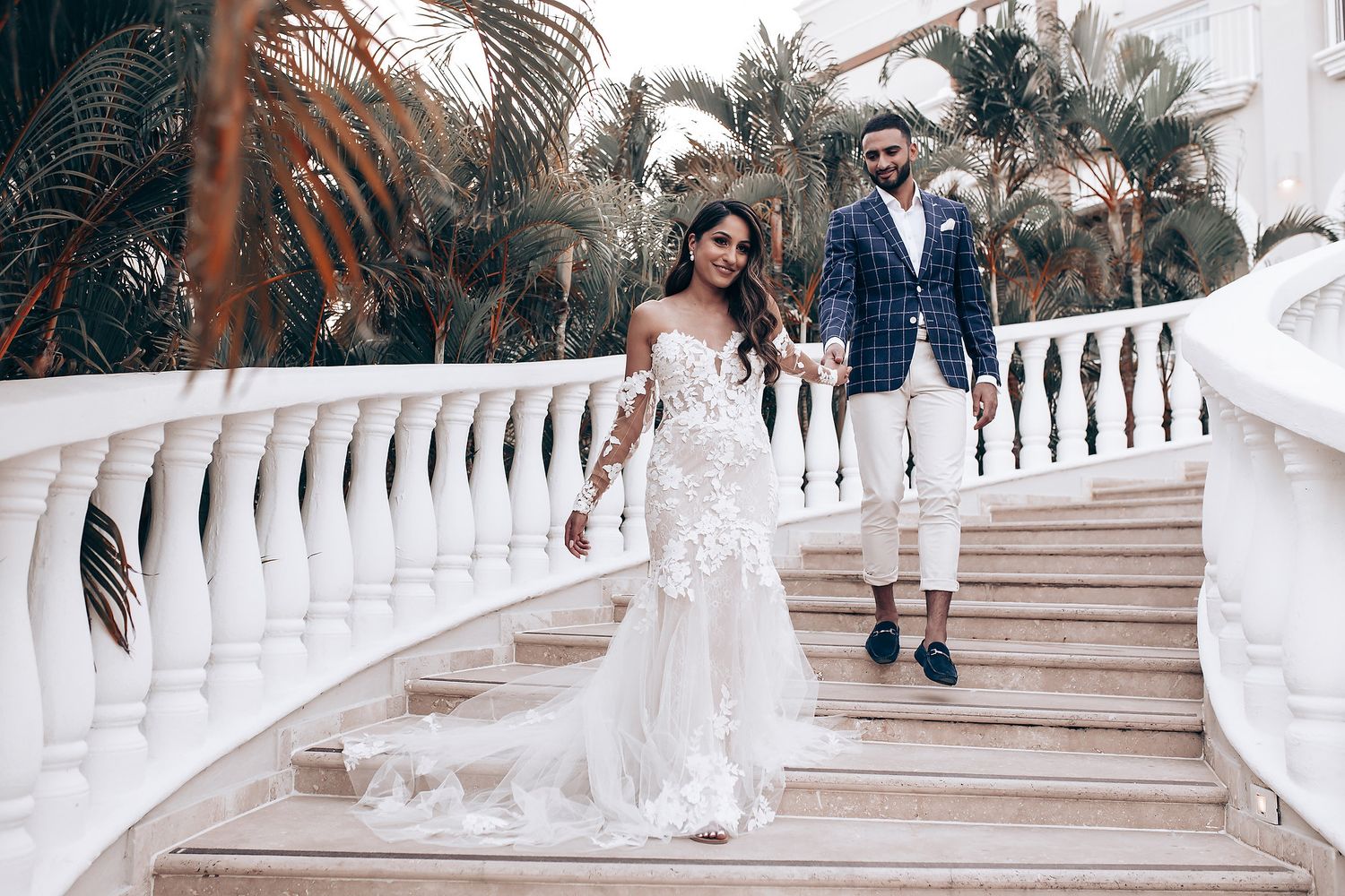 Wedding party poses on grand white staircase with tropical landscaping.