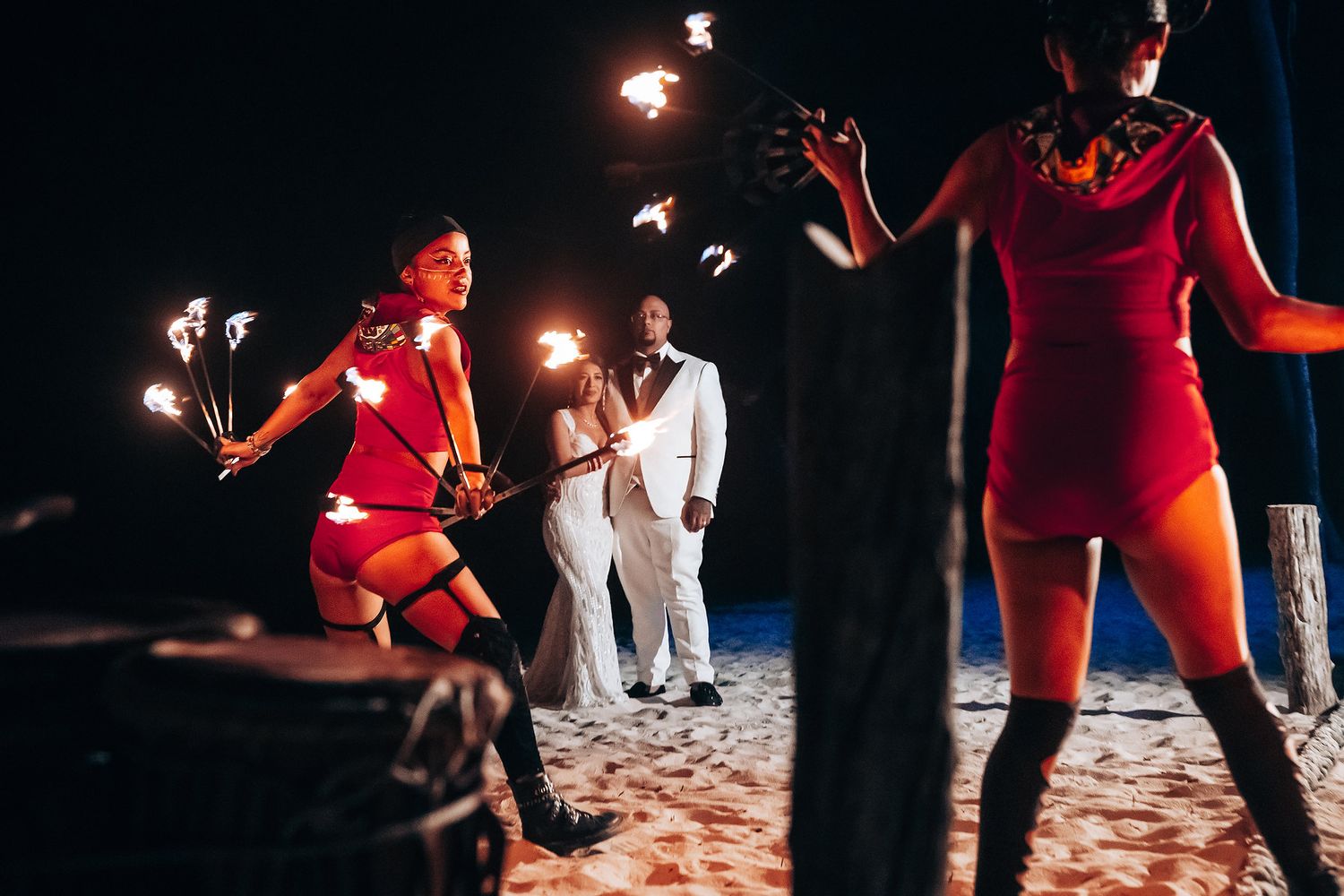 Fire dancers in red costumes perform on the beach during a nighttime wedding celebration.