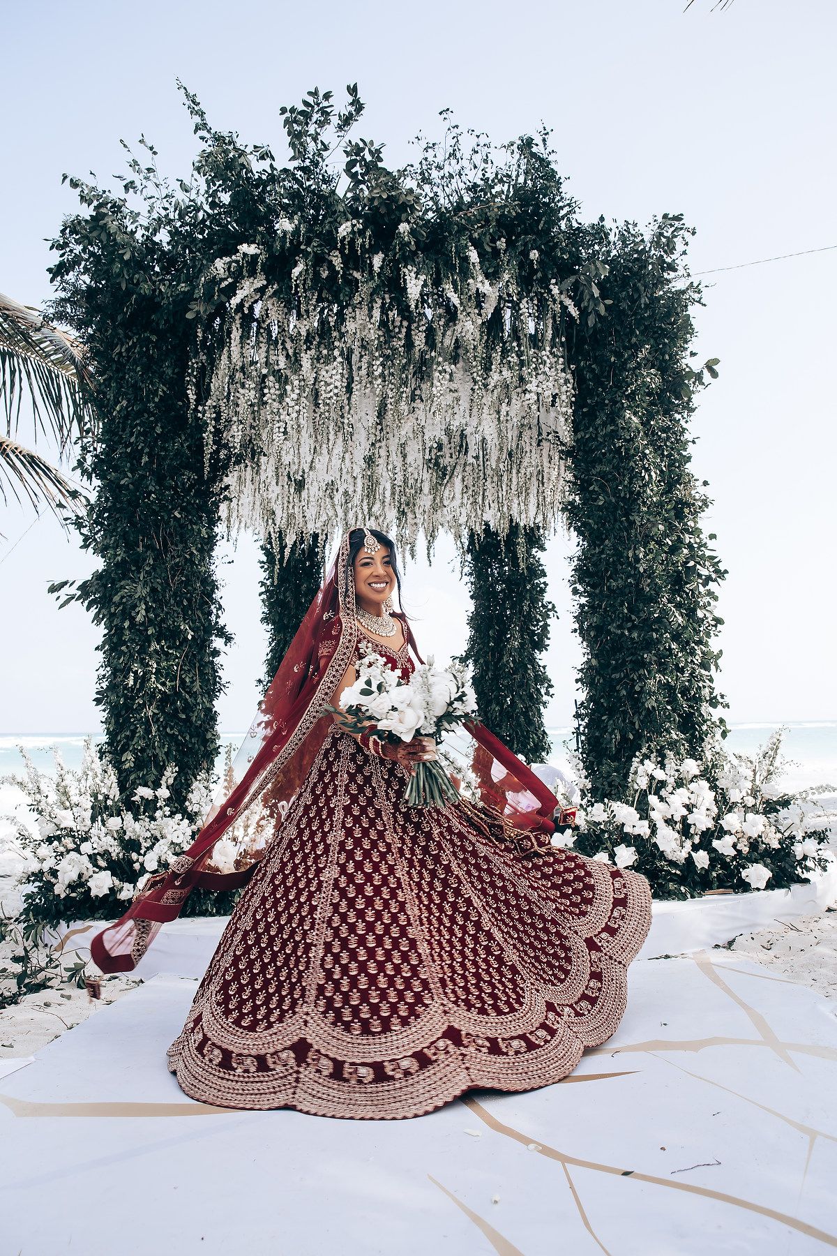 Bride in embellished red lehenga twirls under decorative mandap with hanging florals.