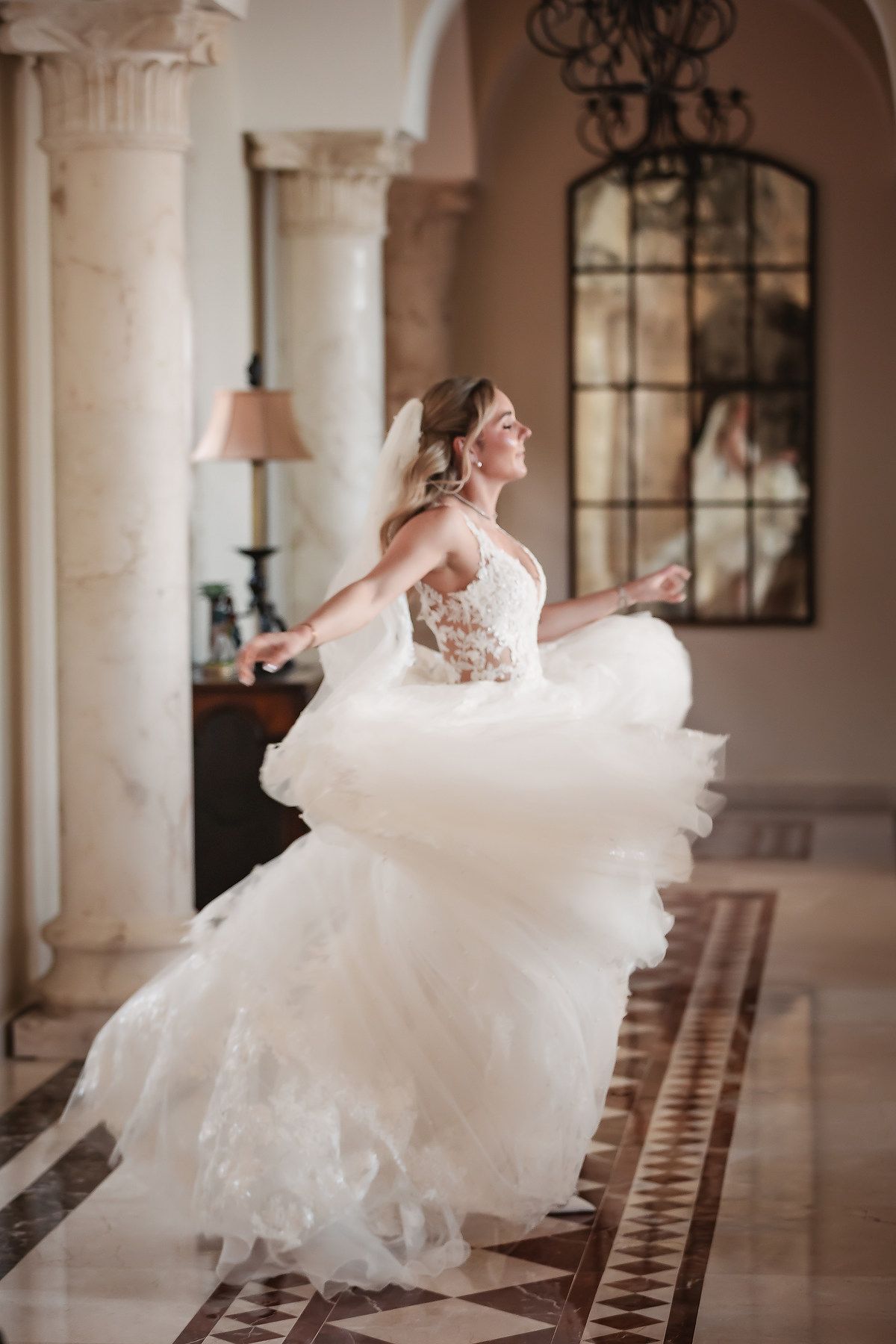 Wedding dress twirls dramatically in an elegant hallway with columns and chandeliers.