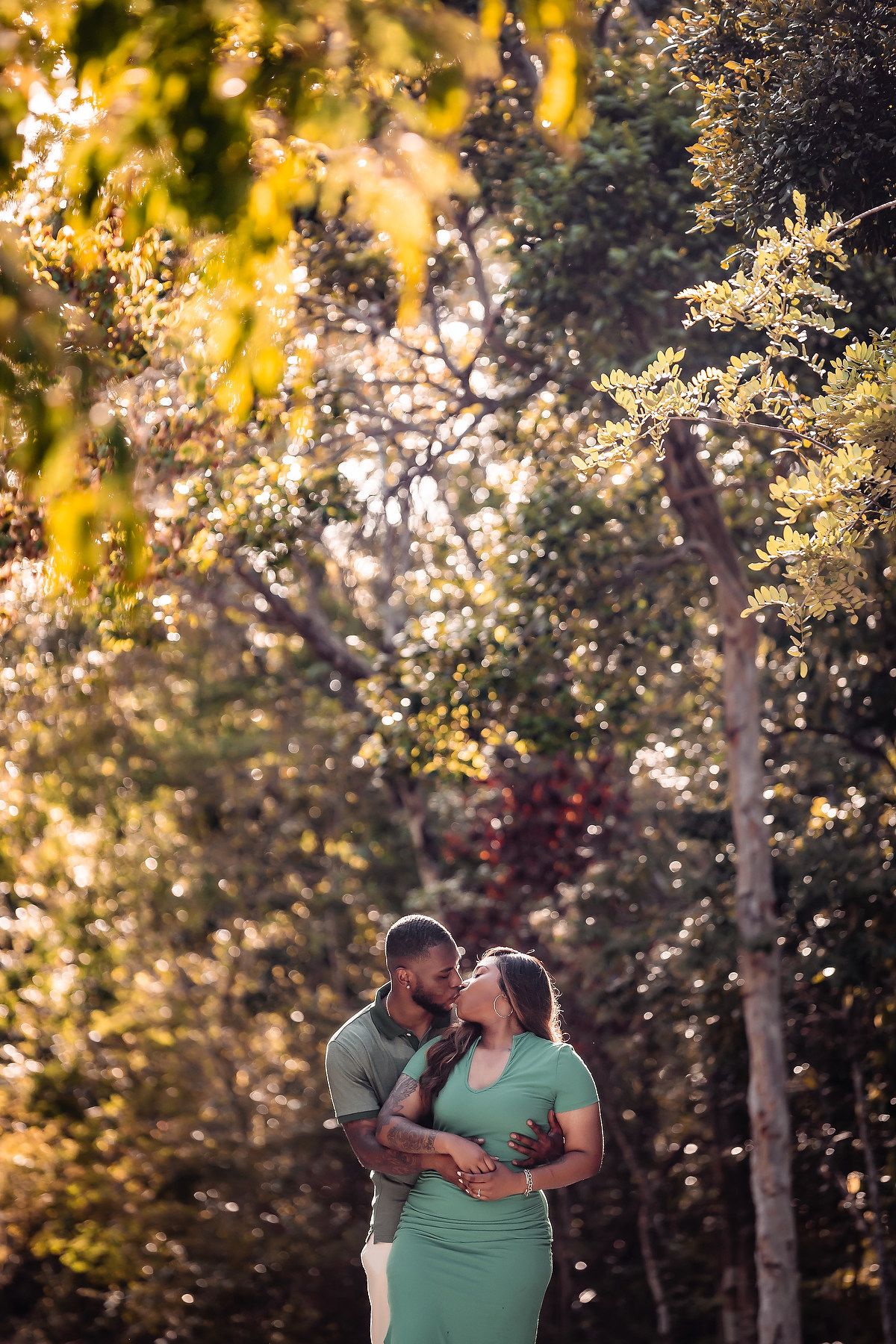 A couple embraces during golden hour in a wooded area with sunlight filtering through trees.