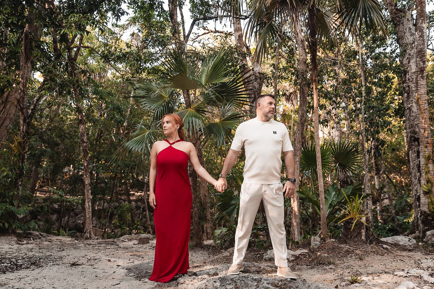 A couple in contrasting red dress and white outfit pose in a lush forest setting.