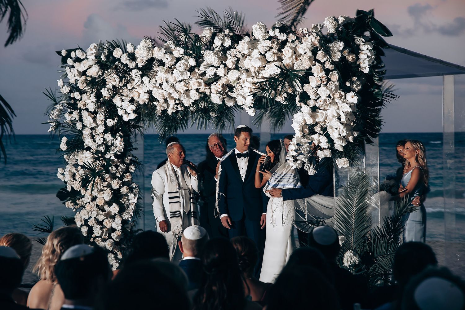 Sunset beach wedding ceremony under an ornate white flower arch.