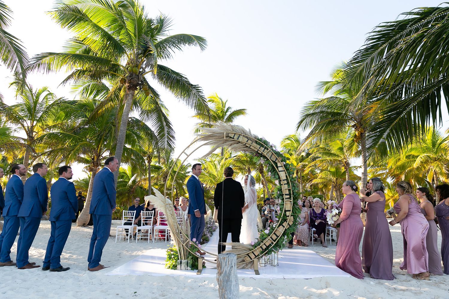 Wedding party members stand in a line formation on a tropical beach with palm trees and ocean views during sunset ceremony.