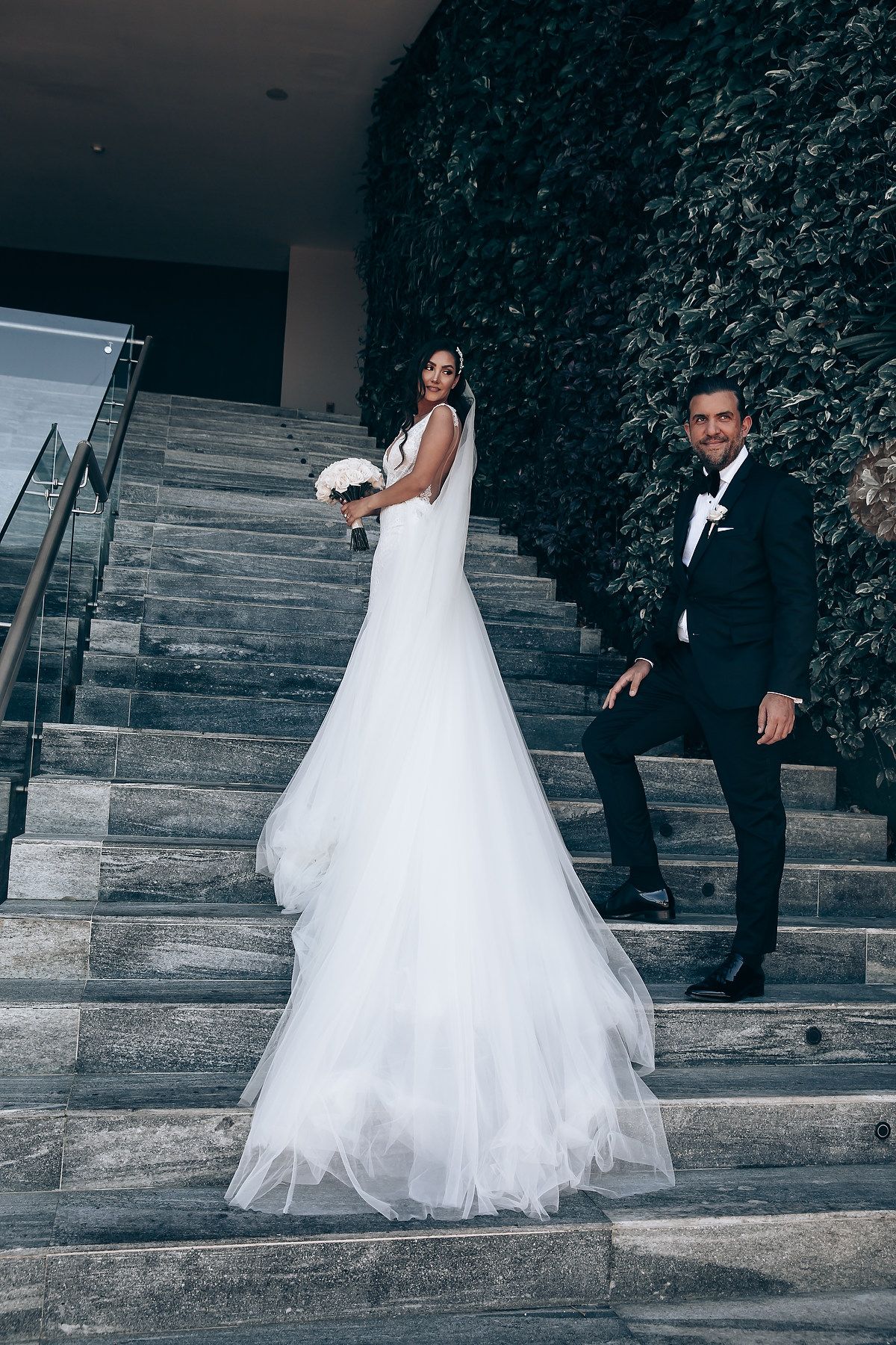 A wedding couple poses on elegant stone steps with a dramatic green wall backdrop in formal attire.