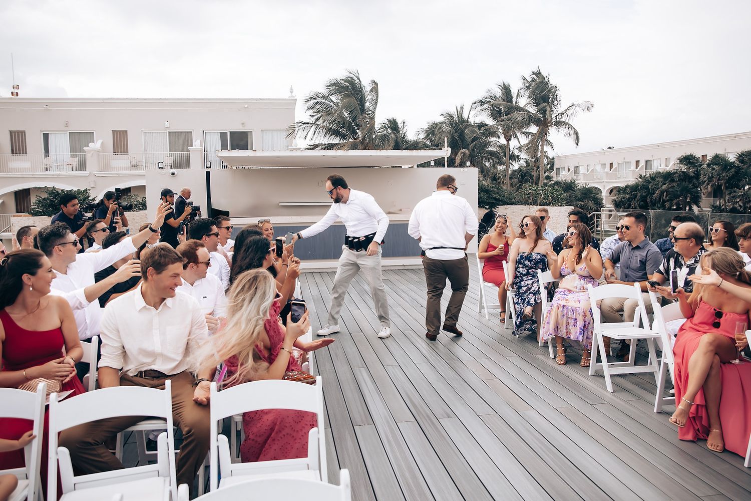 Wedding guests gather on a wooden deck overlooking palm trees at a seaside celebration.