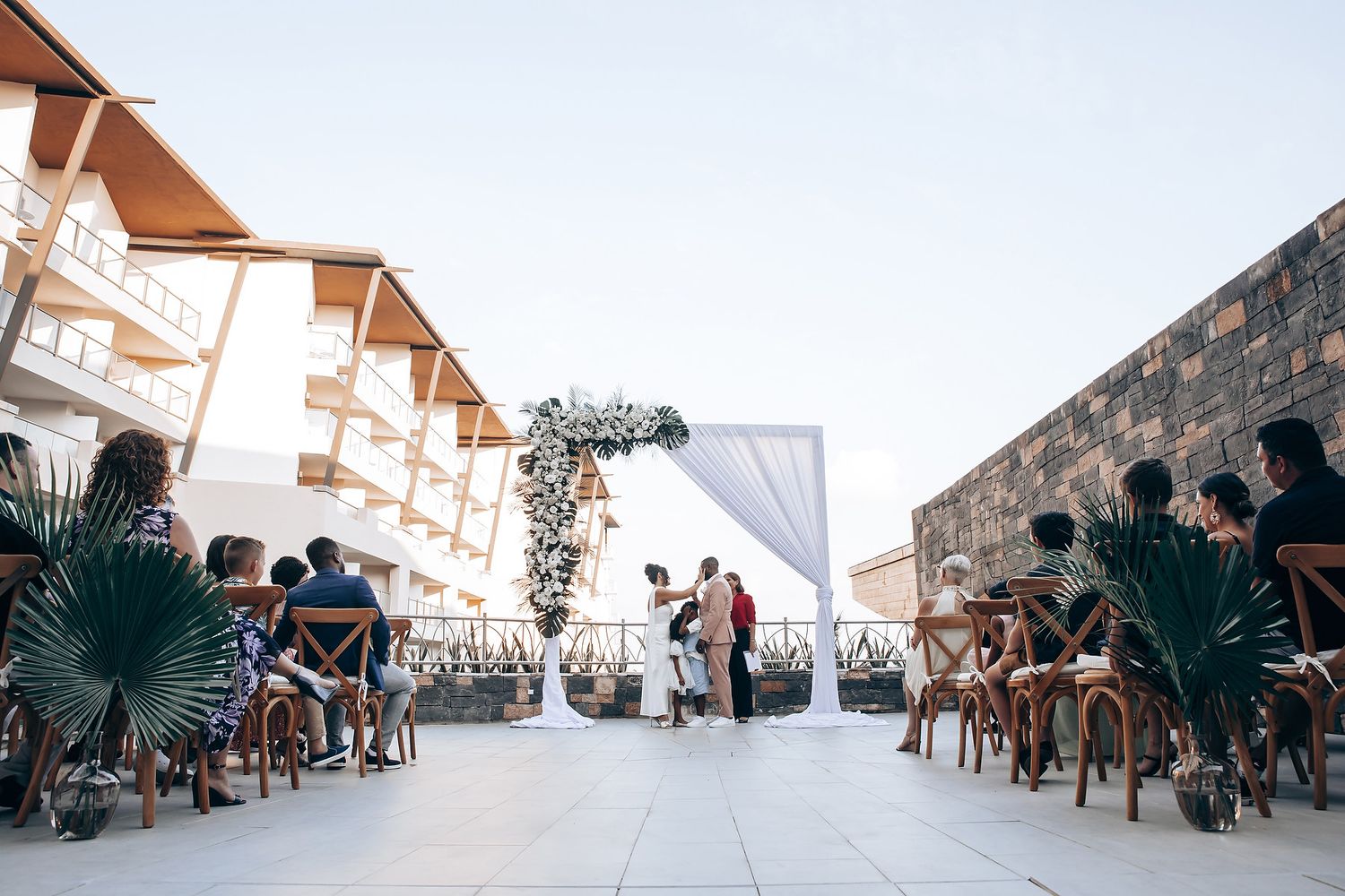 Wedding guests seated in white chairs during an outdoor ceremony with palm trees in the background.