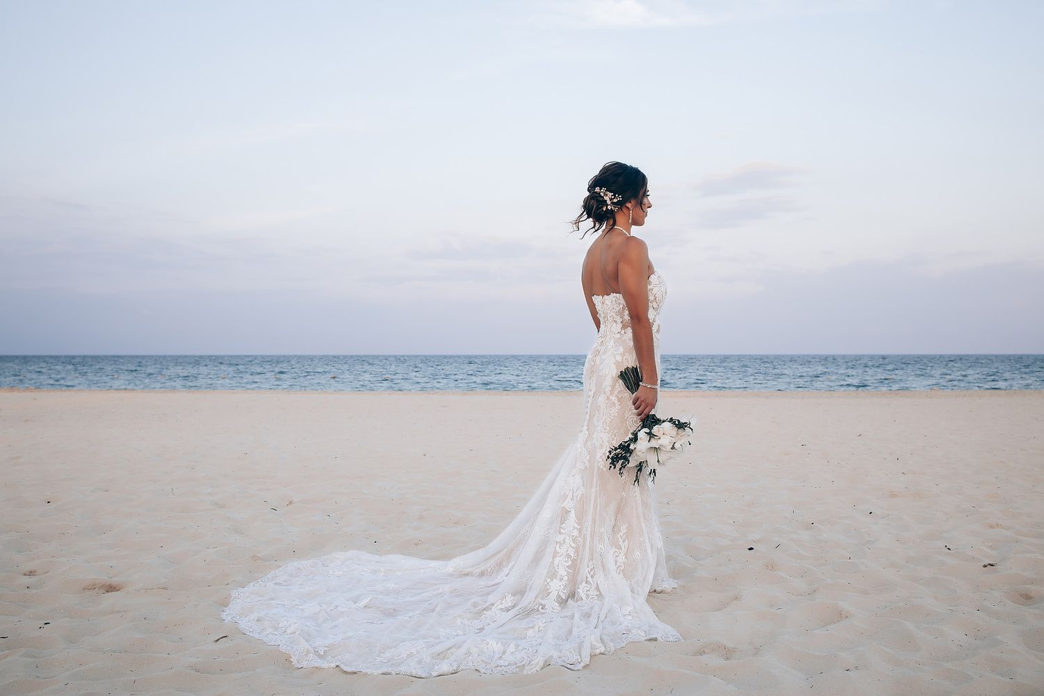 Beautiful white lace wedding dress photographed against sandy beach and ocean backdrop.