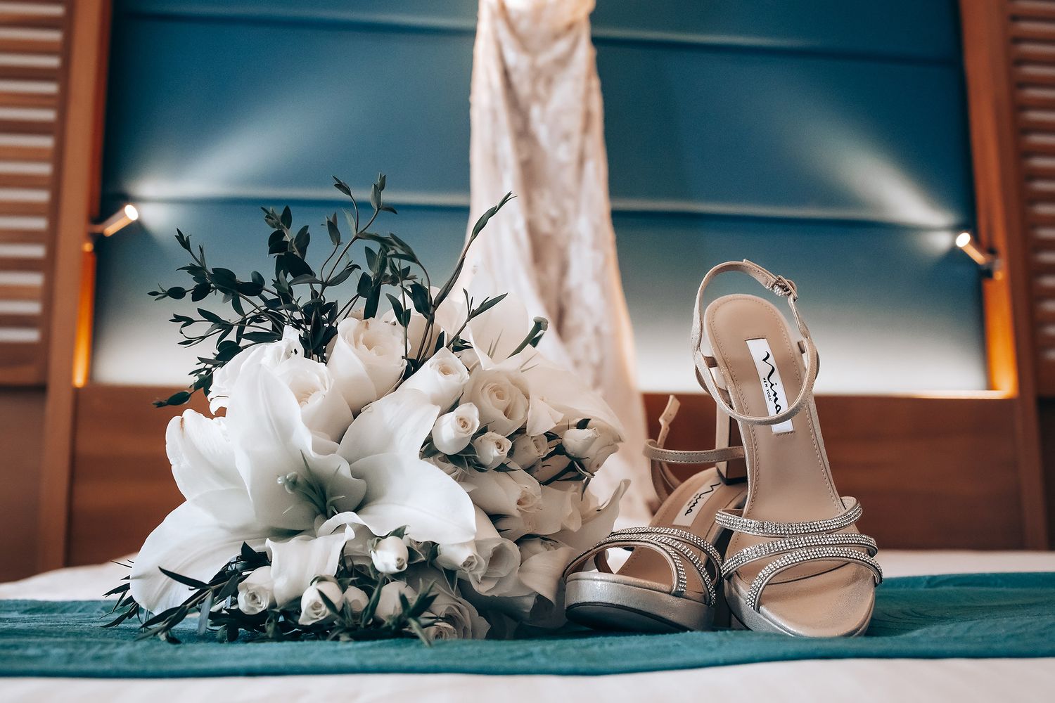 Wedding details featuring elegant silver sandals and a white bridal bouquet with cotton stems on a turquoise surface.