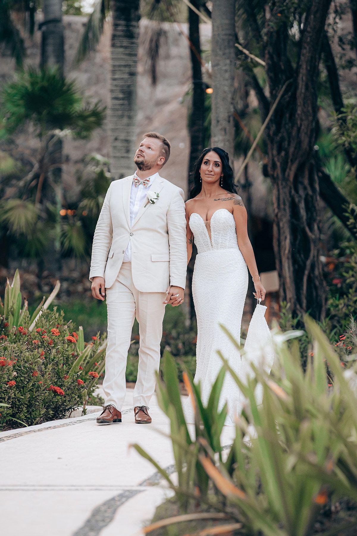 Newlyweds walking along a white pathway surrounded by tropical plants and palm trees.
