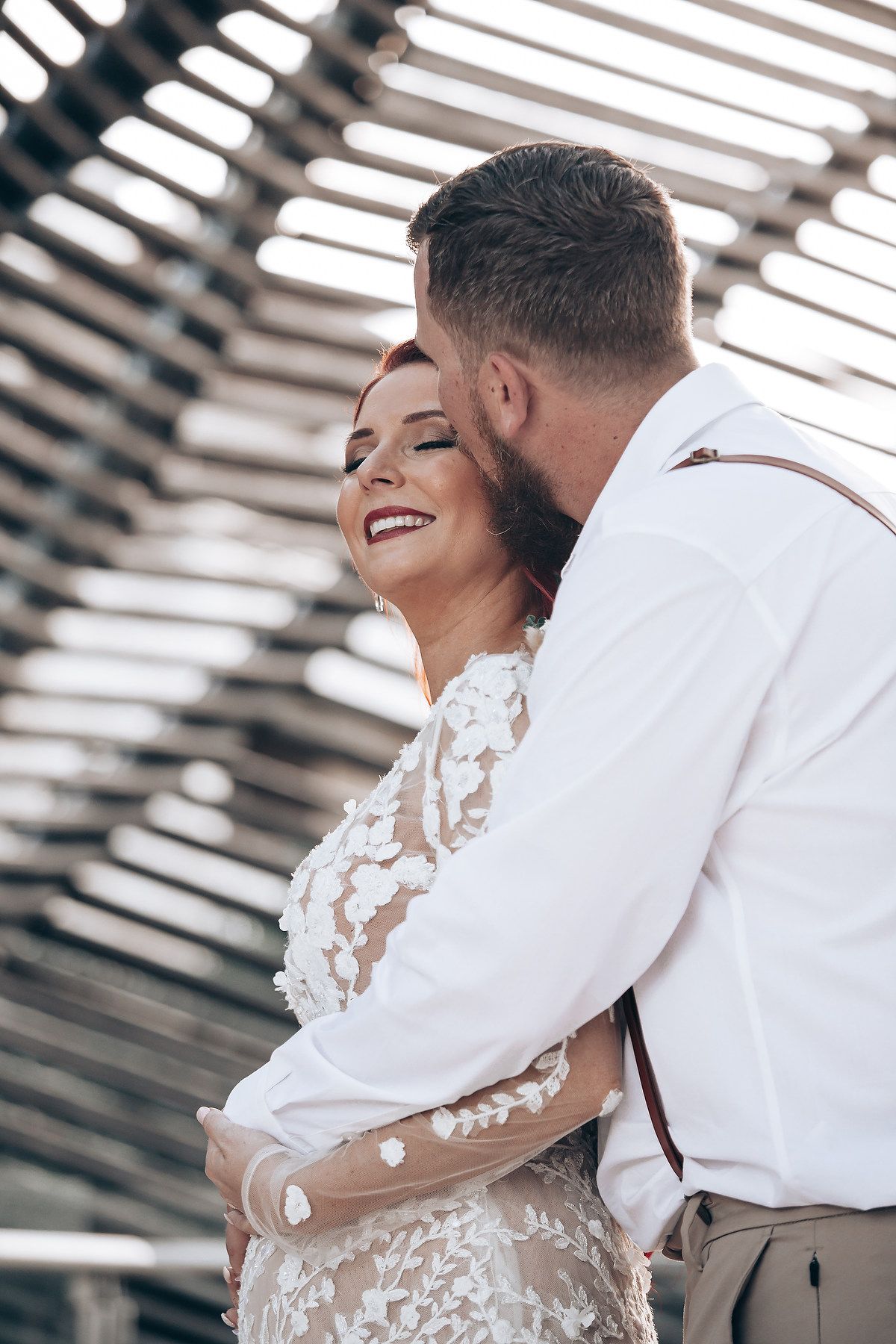 A romantic wedding couple embraces and smiles during an intimate moment captured through window blinds.