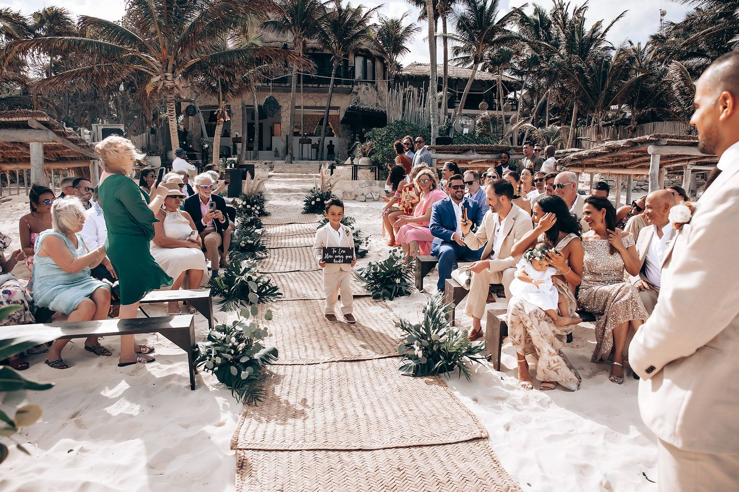Wedding guests seated on benches at outdoor beach ceremony with tropical landscaping.