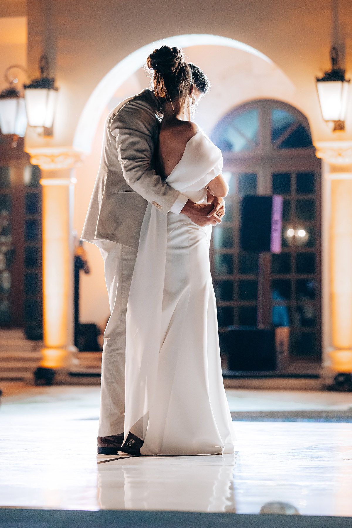 Romantic wedding couple embraces in elegant white attire on a marble floor with arched windows and warm lighting.