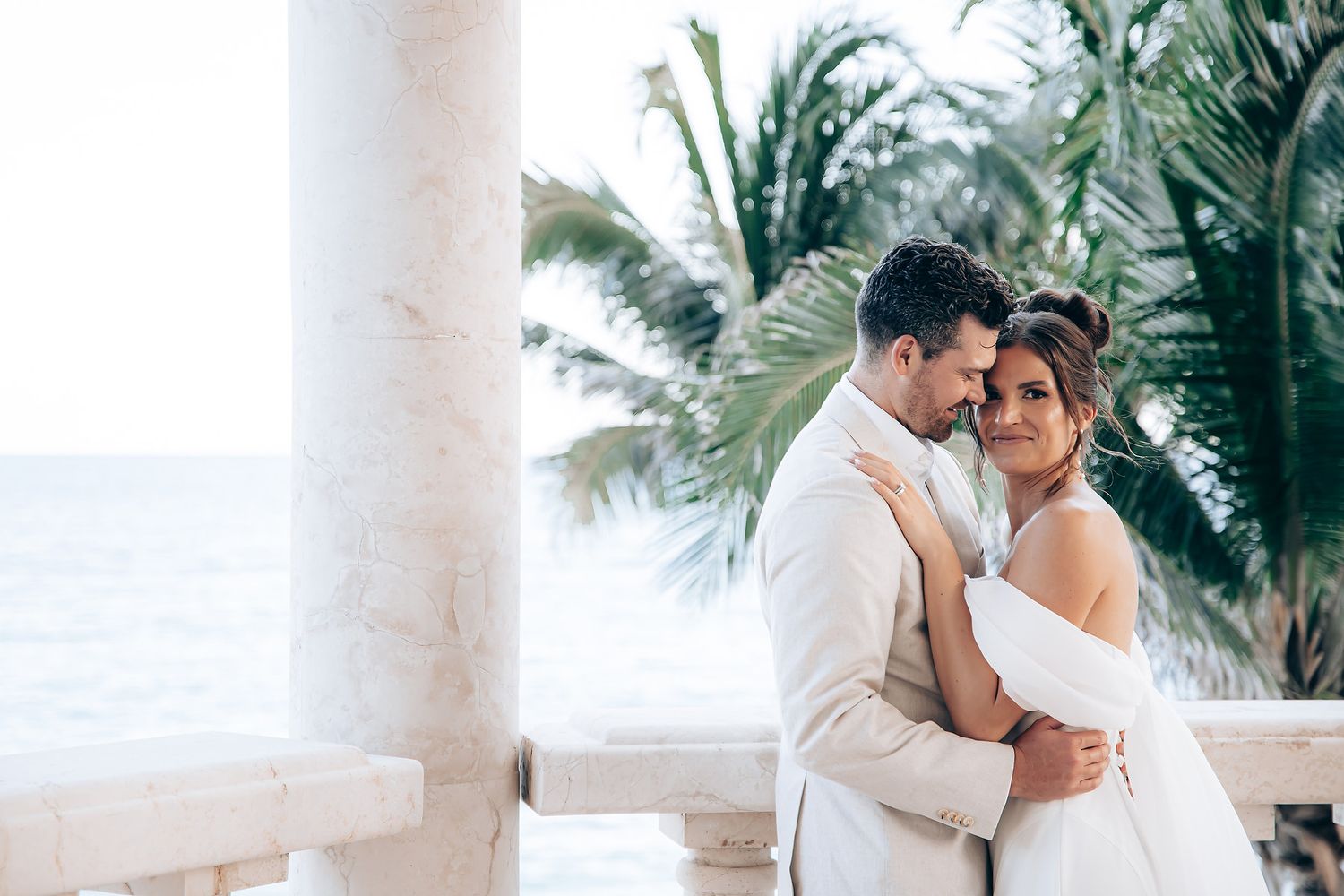 A couple in wedding attire embraces on a tropical beach with palm trees and white columns in the background.
