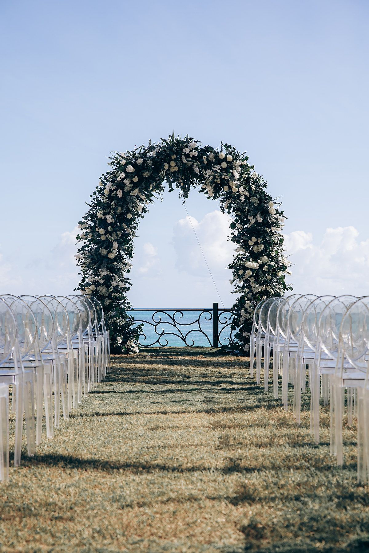 White chairs line up symmetrically leading to a floral arch for an outdoor wedding ceremony setup in a grassy field.