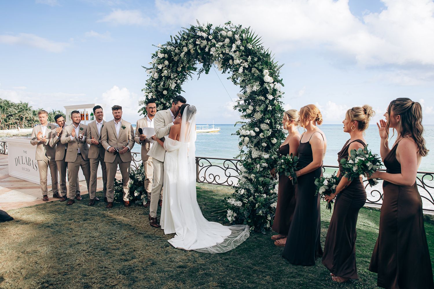 Wedding party gathered outdoors under floral arch with scenic waterfront backdrop during sunset ceremony.
