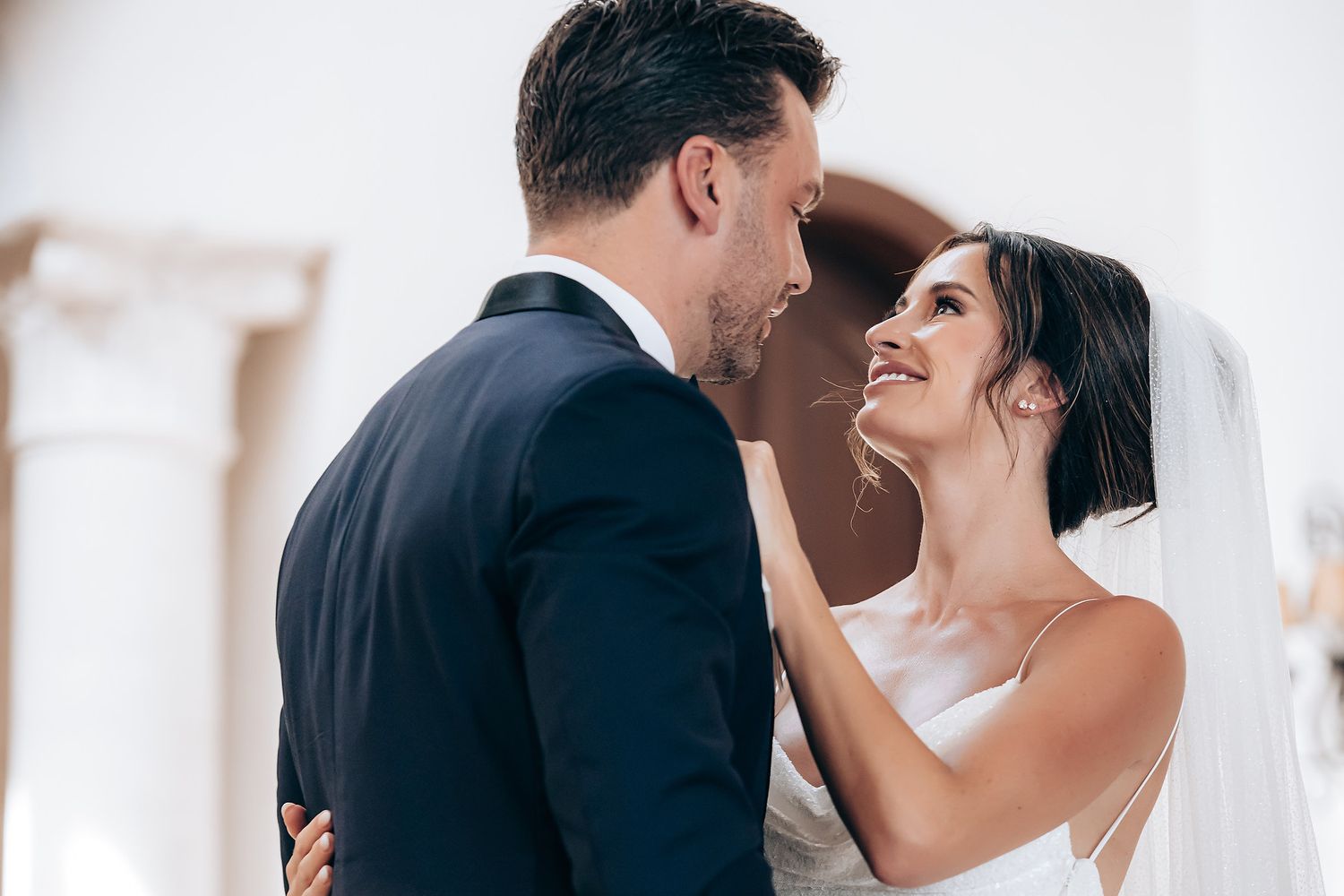 A romantic wedding couple shares an intimate moment while dancing together in an elegant white venue.