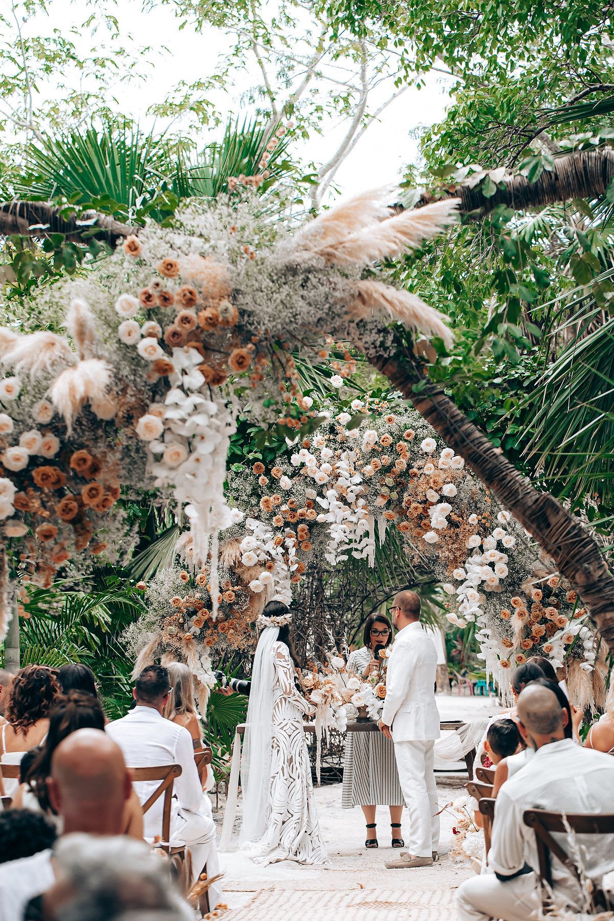 A romantic outdoor wedding ceremony setup with a floral arch featuring blush and white blooms under tropical palm trees.