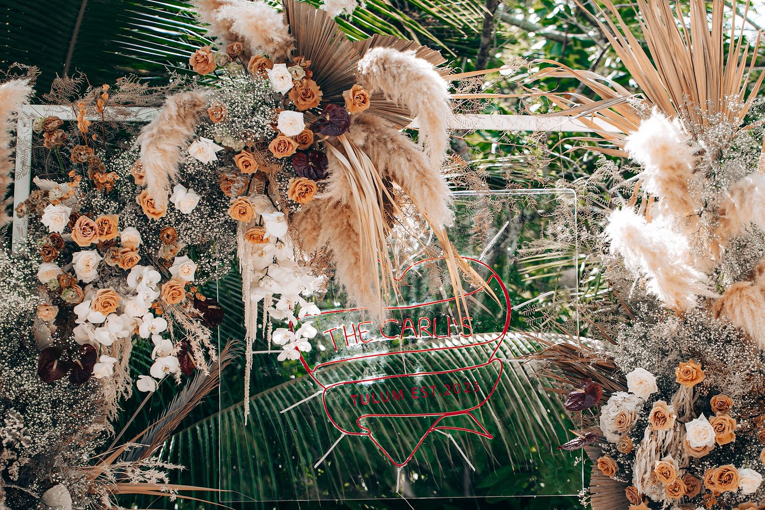 Elegant wedding arch decorated with dried pampas grass, white roses and palm fronds in a tropical outdoor setting.
