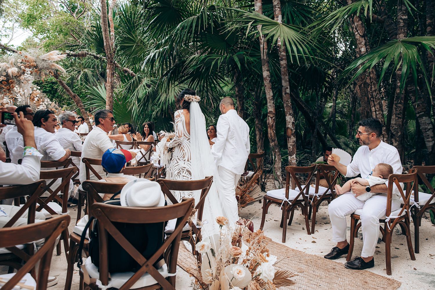 Outdoor wedding ceremony in a tropical garden setting with wooden cross-back chairs and palm trees.