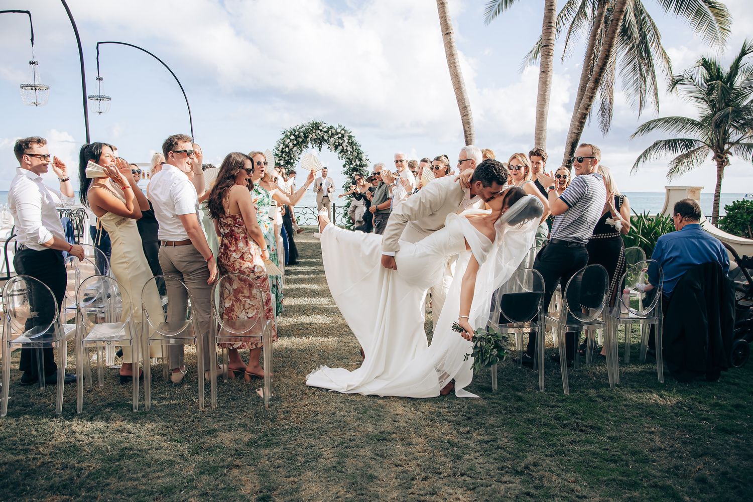 Wedding ceremony on a tropical beach with palm trees and guests seated on clear chairs around a floral arch.