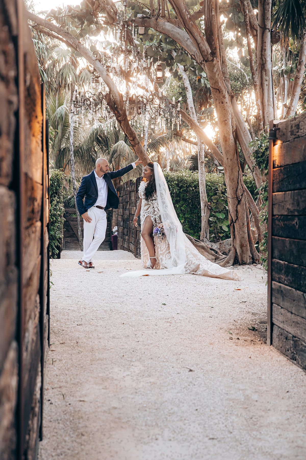 A couple in wedding attire poses for romantic photos in a garden setting with tall trees.