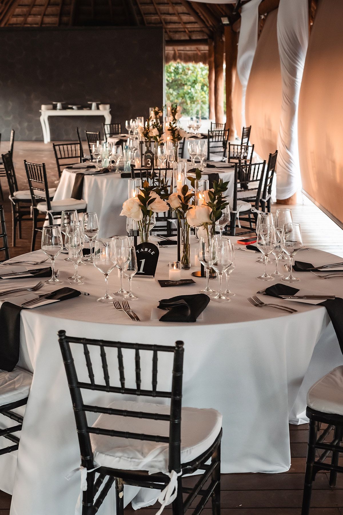 Wedding reception tables set with white linens and black chiavari chairs under rustic wooden beams.