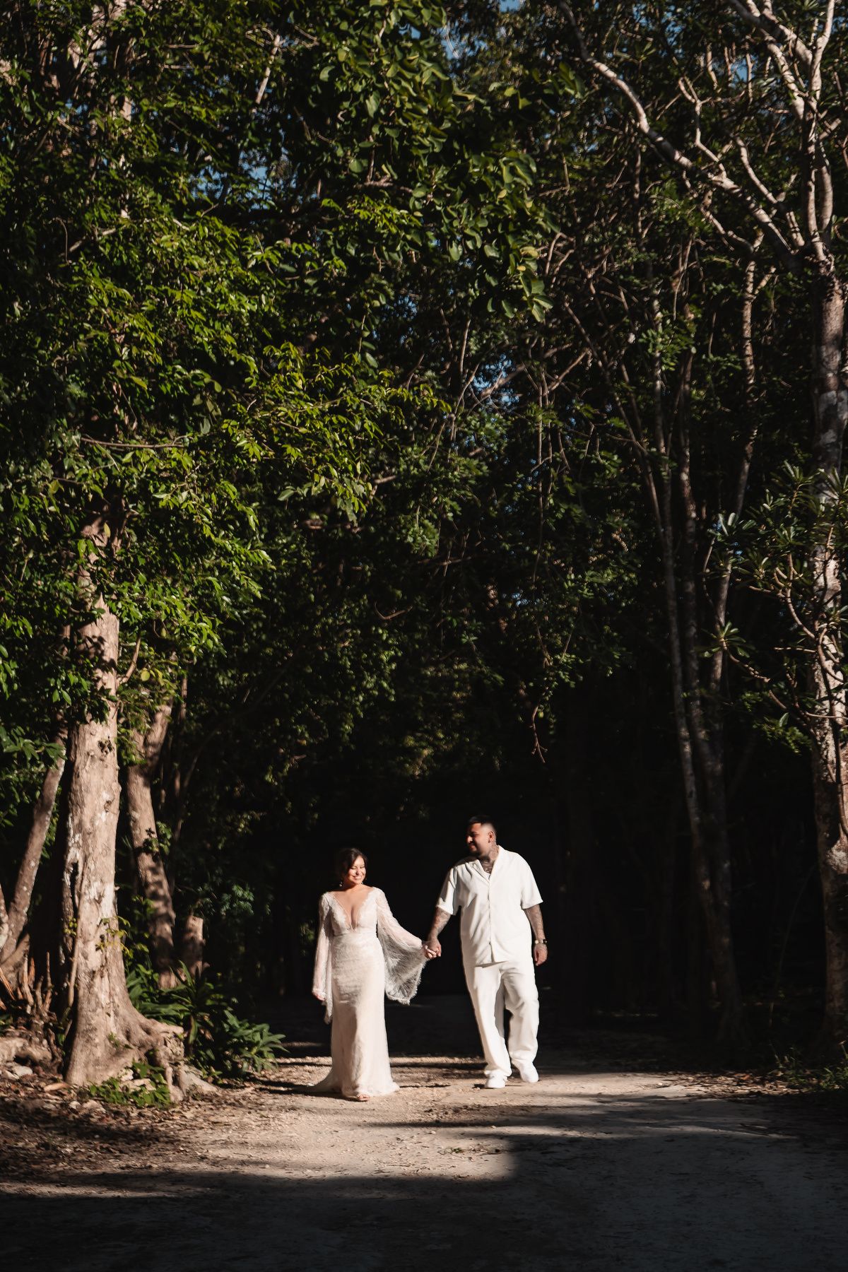 A couple walking hand in hand through a sunlit forest path in wedding attire.