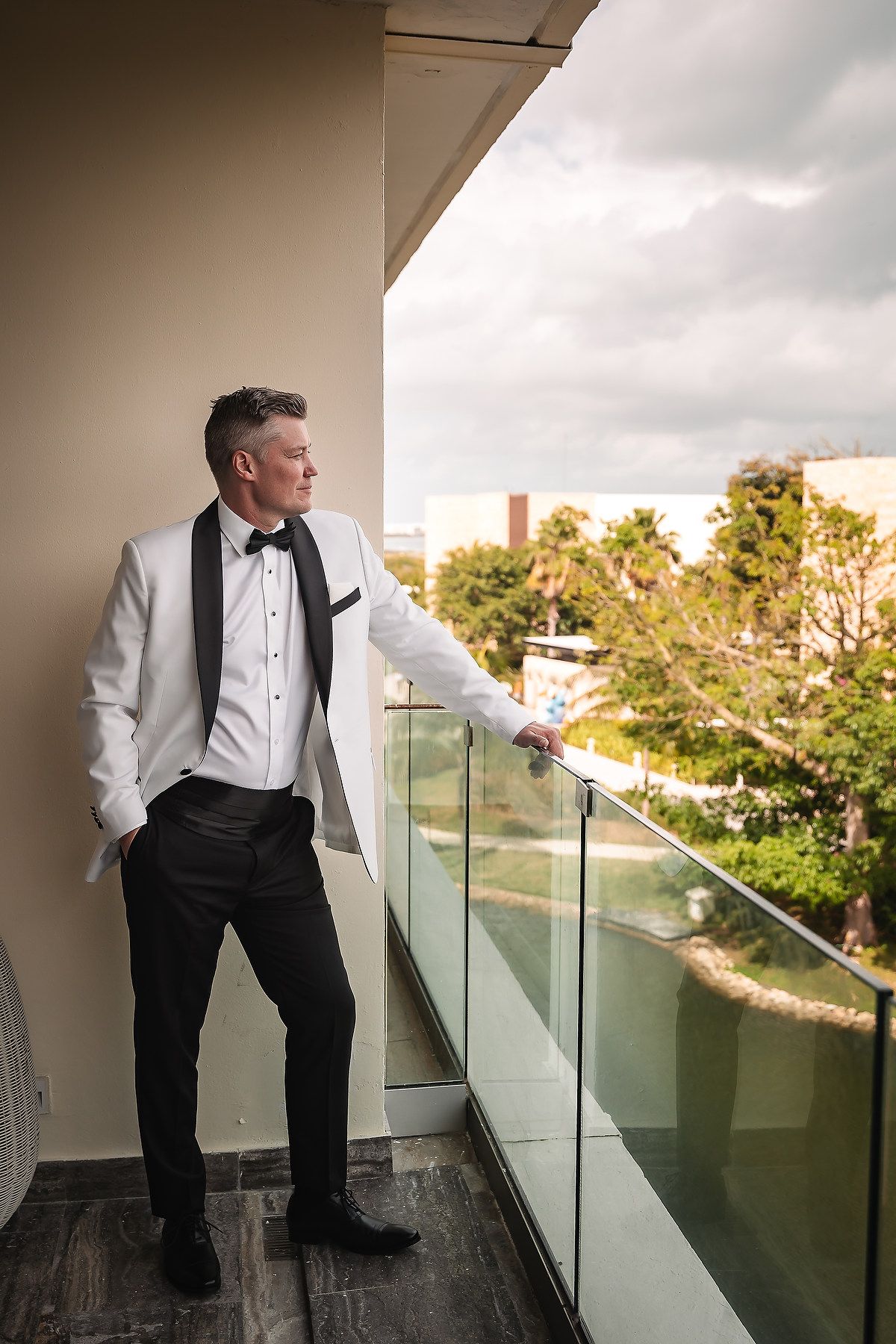 A person in formal black and white attire stands on a balcony overlooking palm trees at sunset.