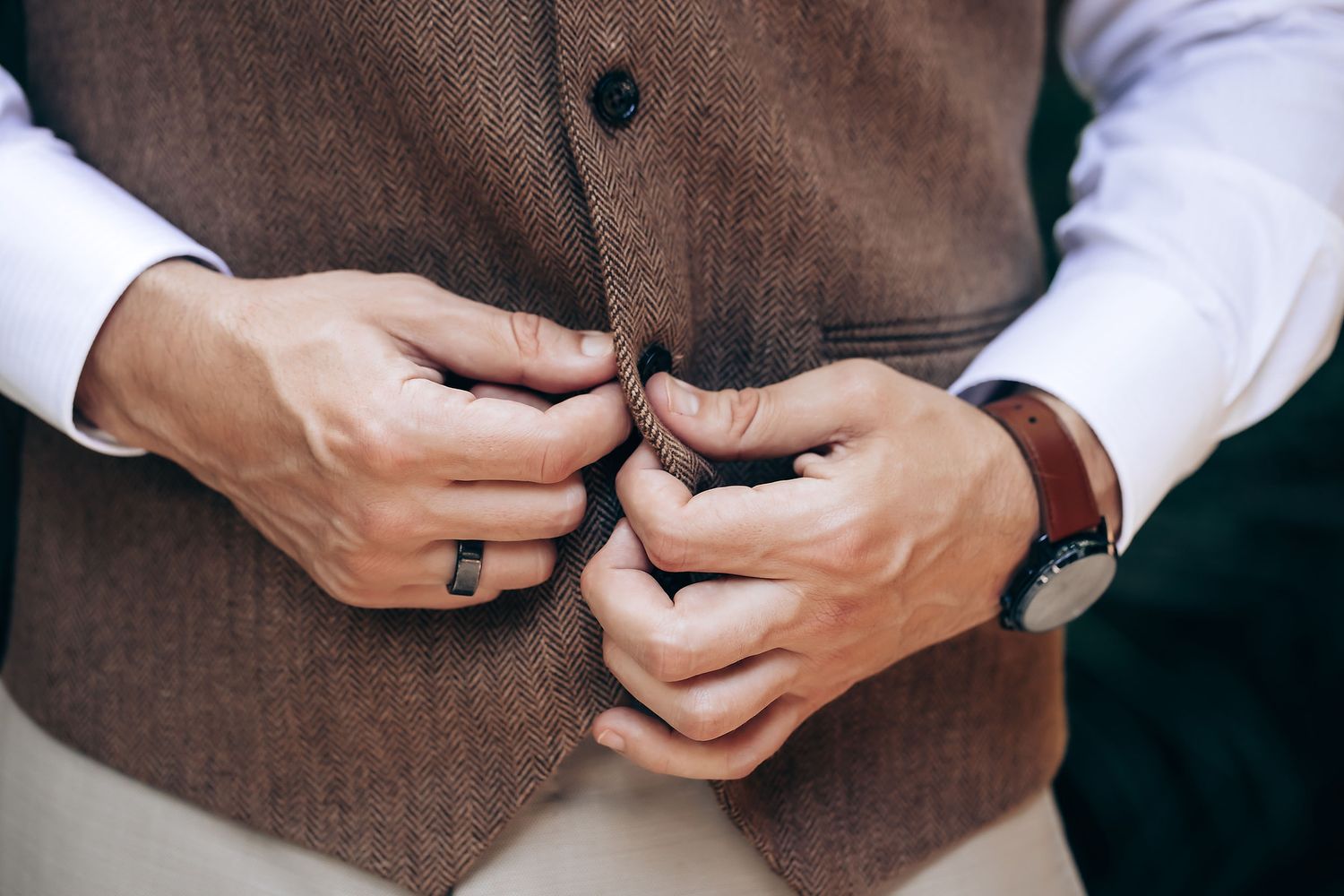 Close-up of hands buttoning a brown tweed vest with a watch visible on the wrist.