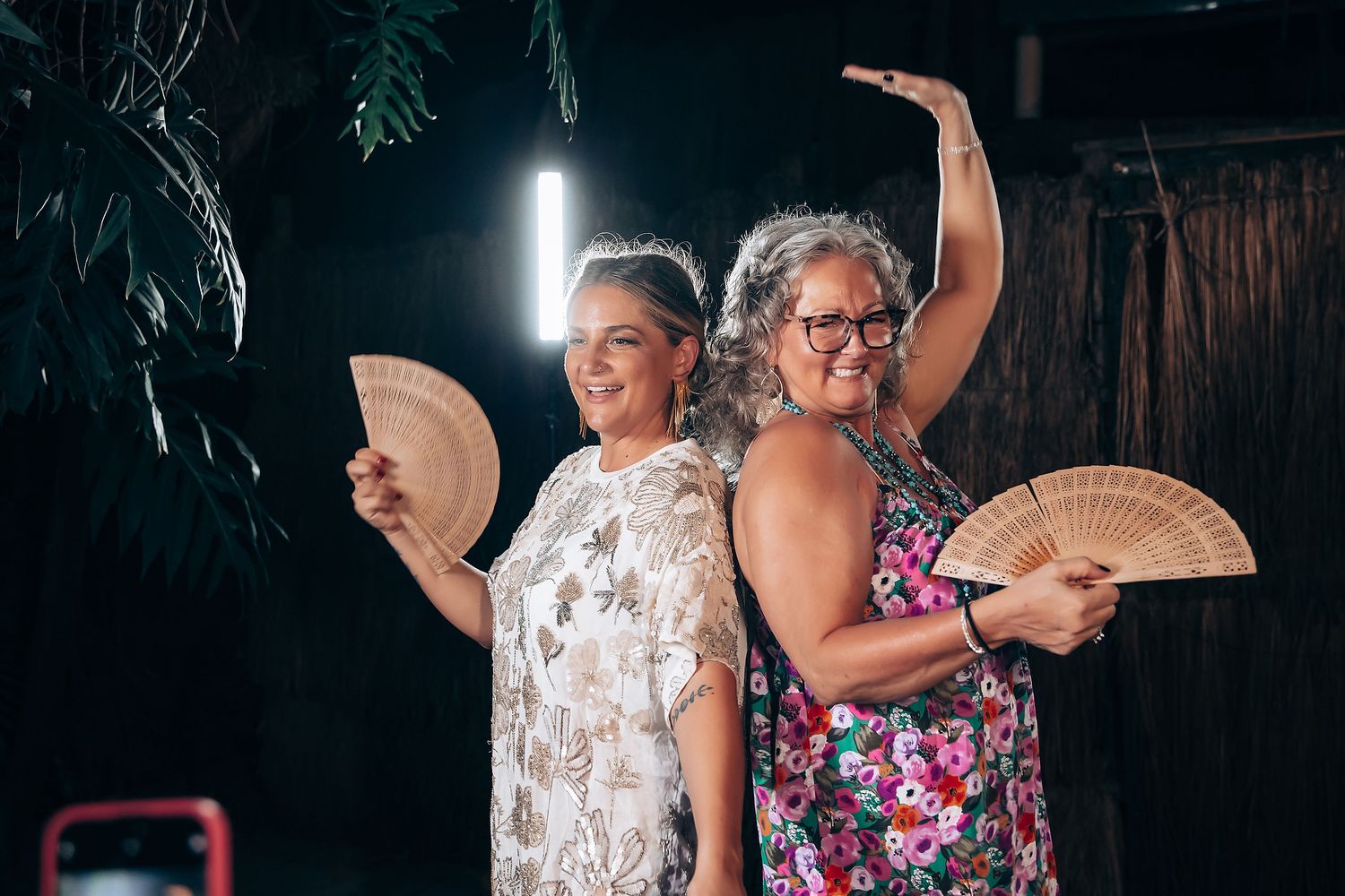 Two women in summer dresses pose back-to-back holding decorative hand fans at an evening event.