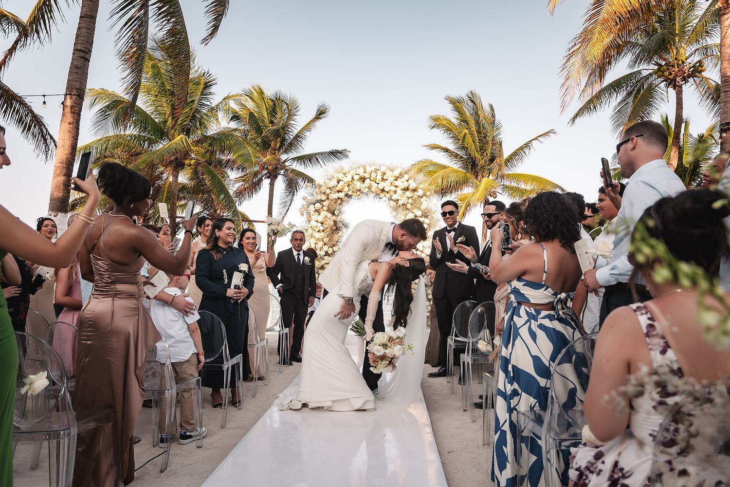 Beachside wedding celebration with guests gathered around a white aisle runner under swaying palm trees.